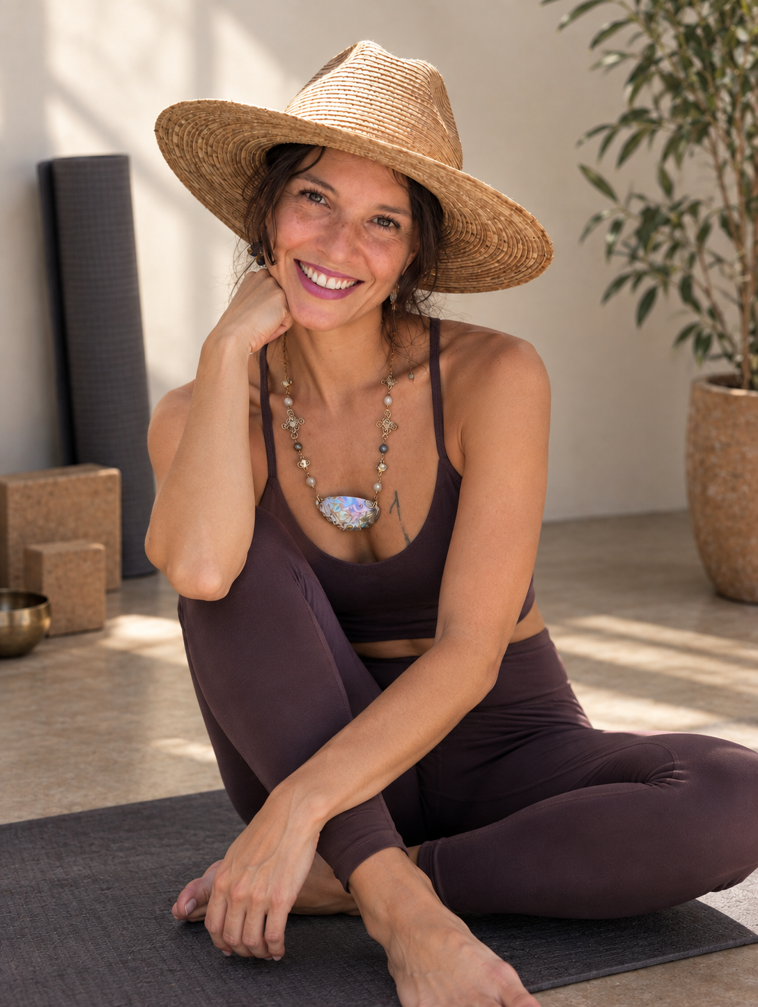 Karina Cordoba smiling warmly in a straw hat, seated on her yoga mat